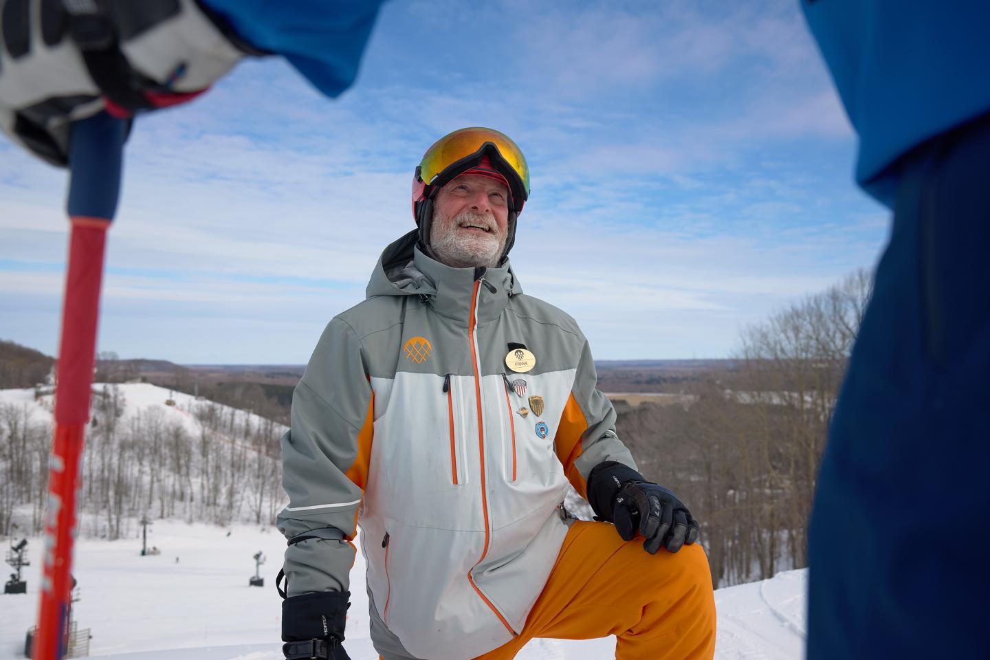 Man in ski gear kneels on snow, smiling under a clear blue sky.