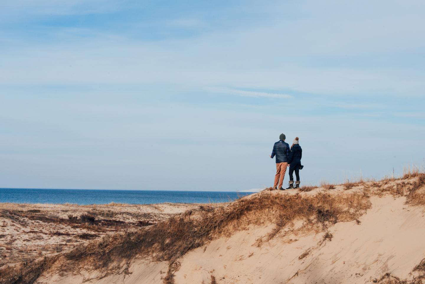 Two people stand on a sandy hill overlooking the sea under a blue sky.