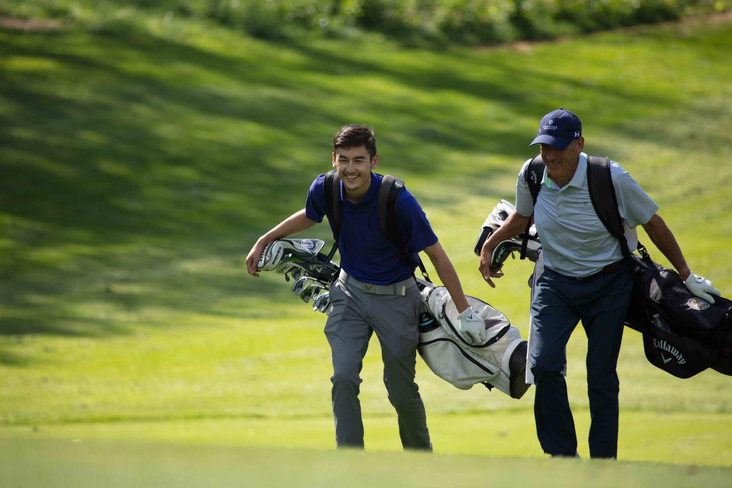 Two golfers walking on a sunny golf course, carrying bags and smiling.
