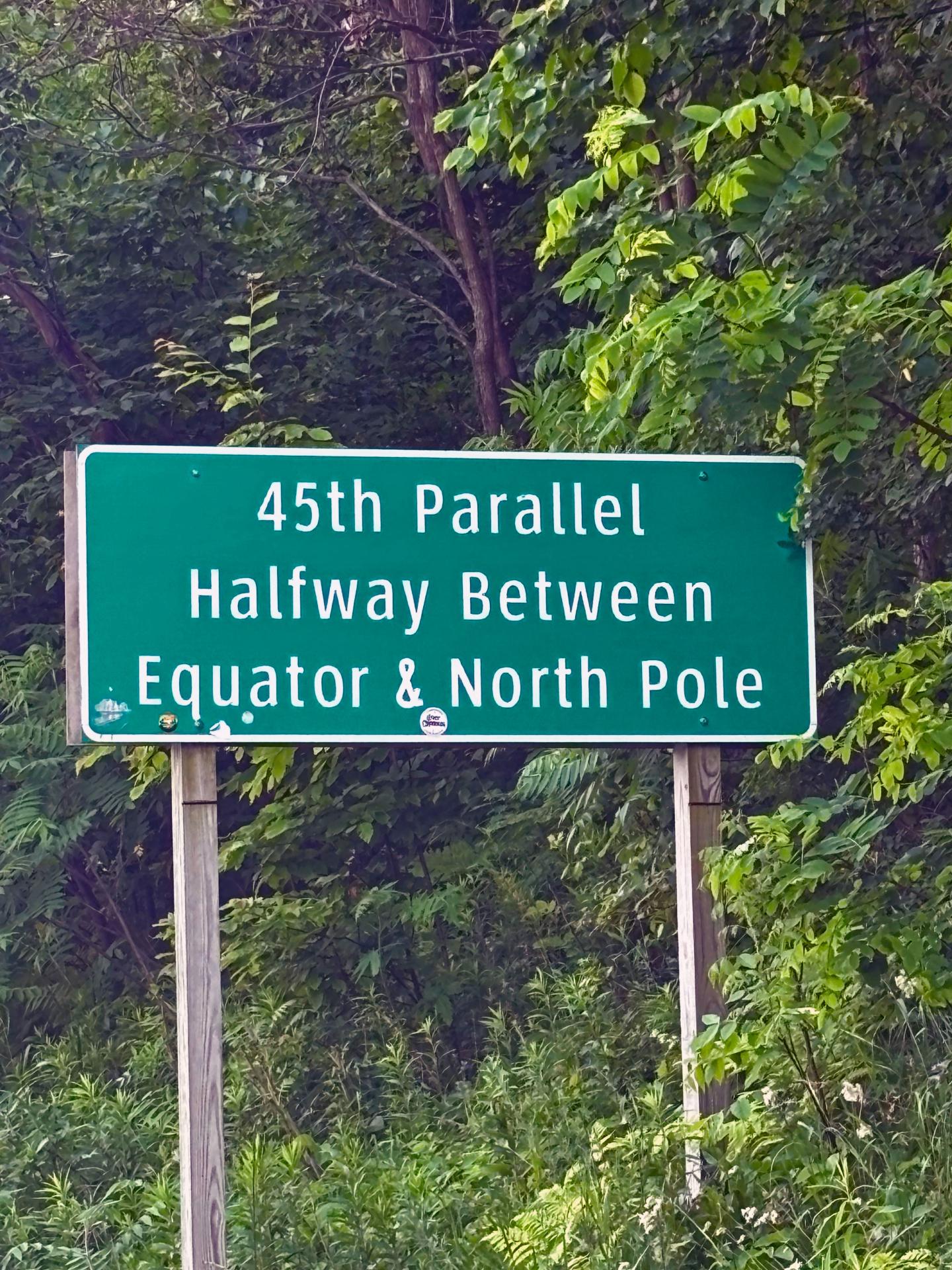 Green sign reading "45th Parallel, Halfway Between Equator & North Pole" amidst trees.