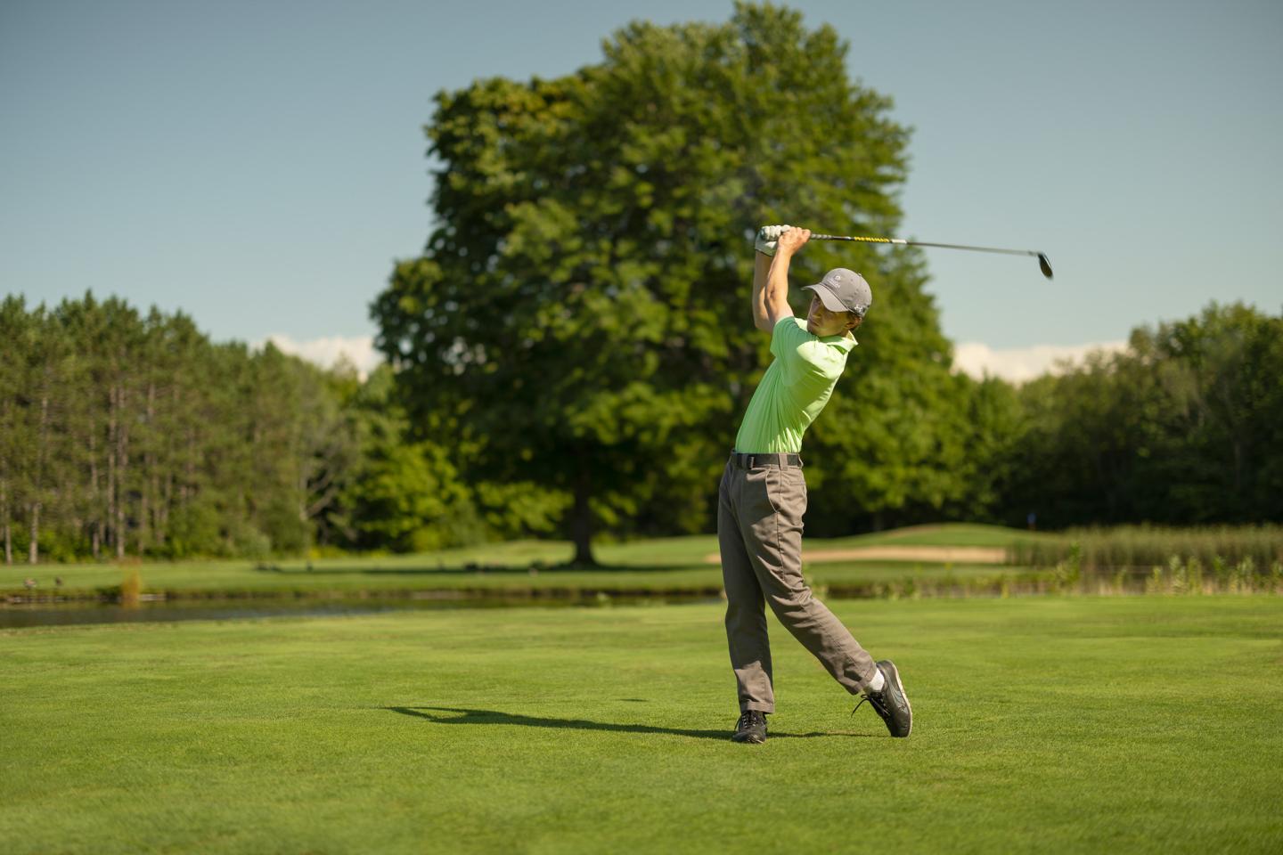 Golfer swinging club on sunny day, green fairway, trees in background.