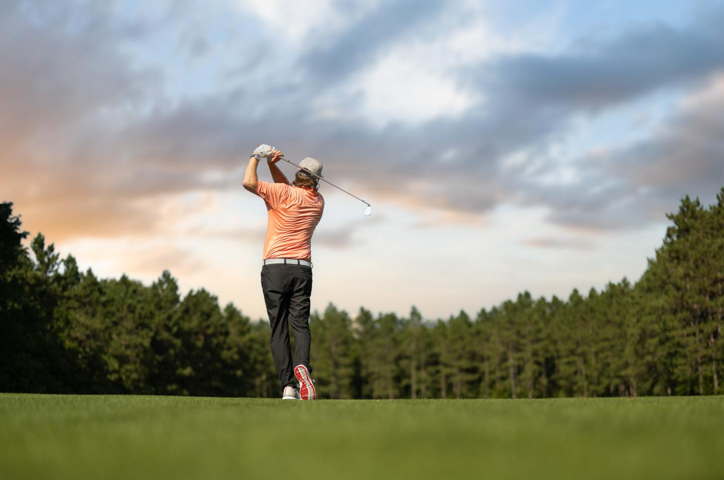 Golfer swinging club on green, trees and sunset sky in the background.