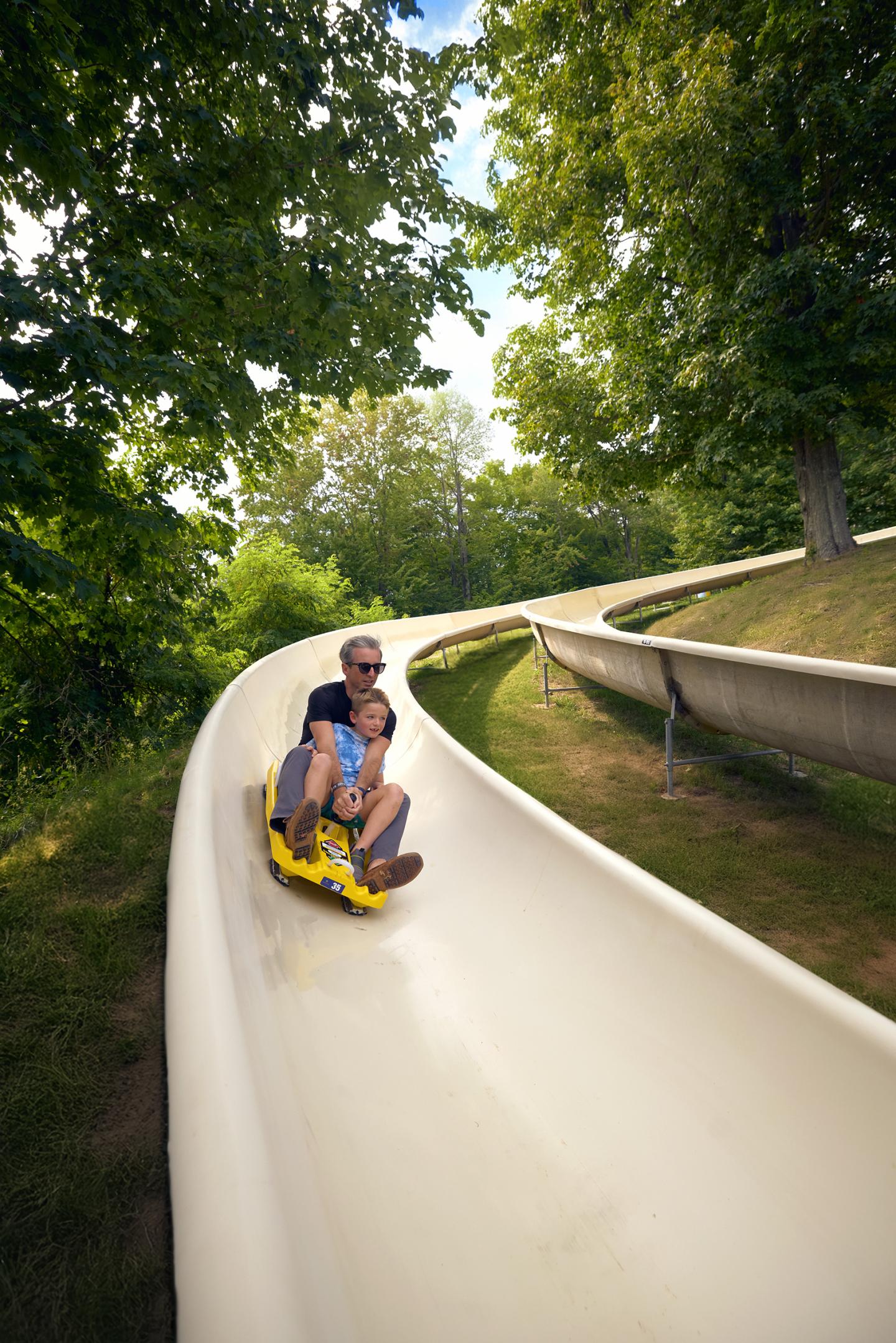 Father and child sliding down a curved slide surrounded by trees.