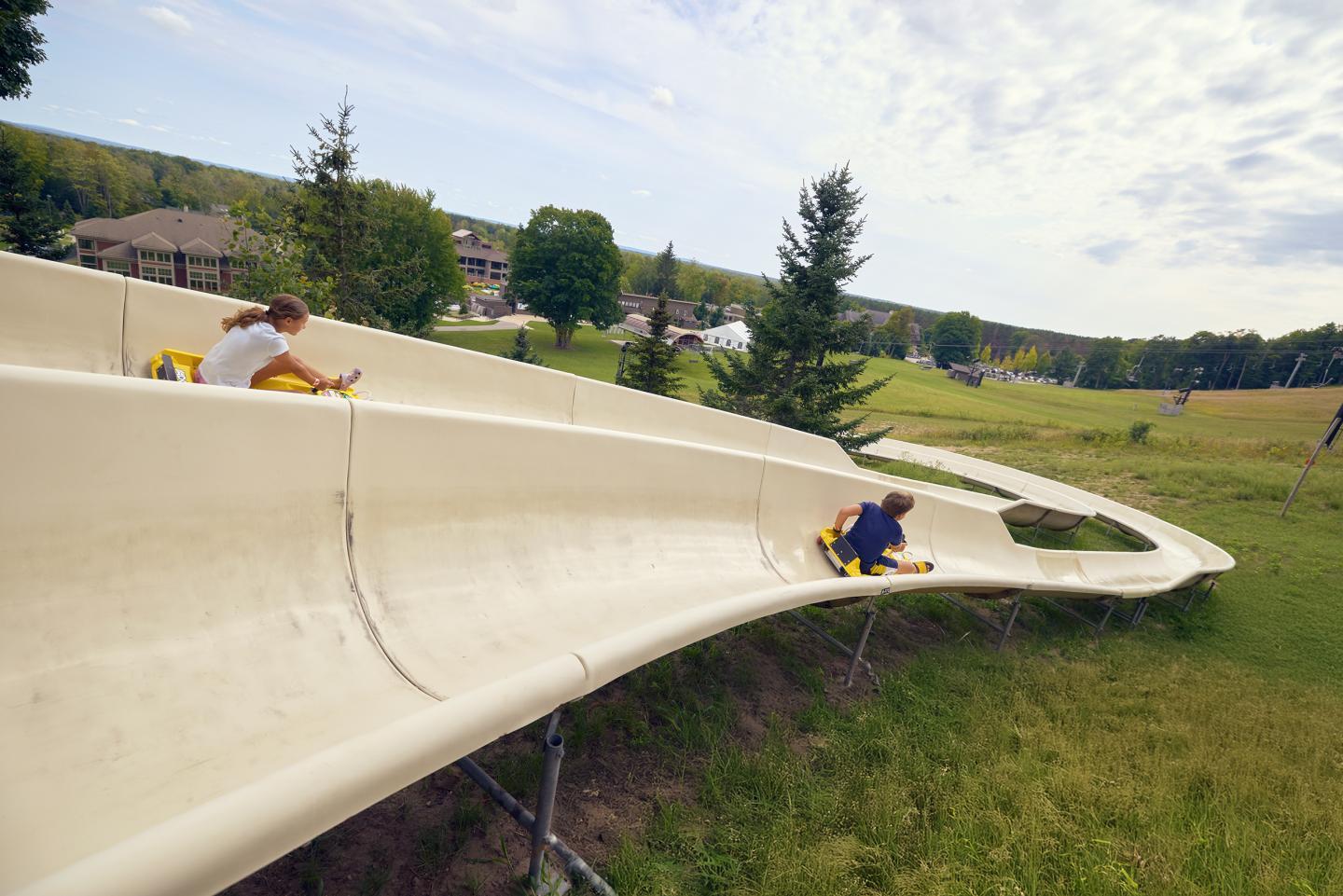 Two people ride down a curved outdoor slide on grass.