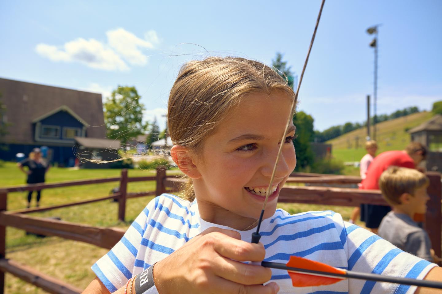 Child smiling while aiming an arrow outdoors under a clear sky.