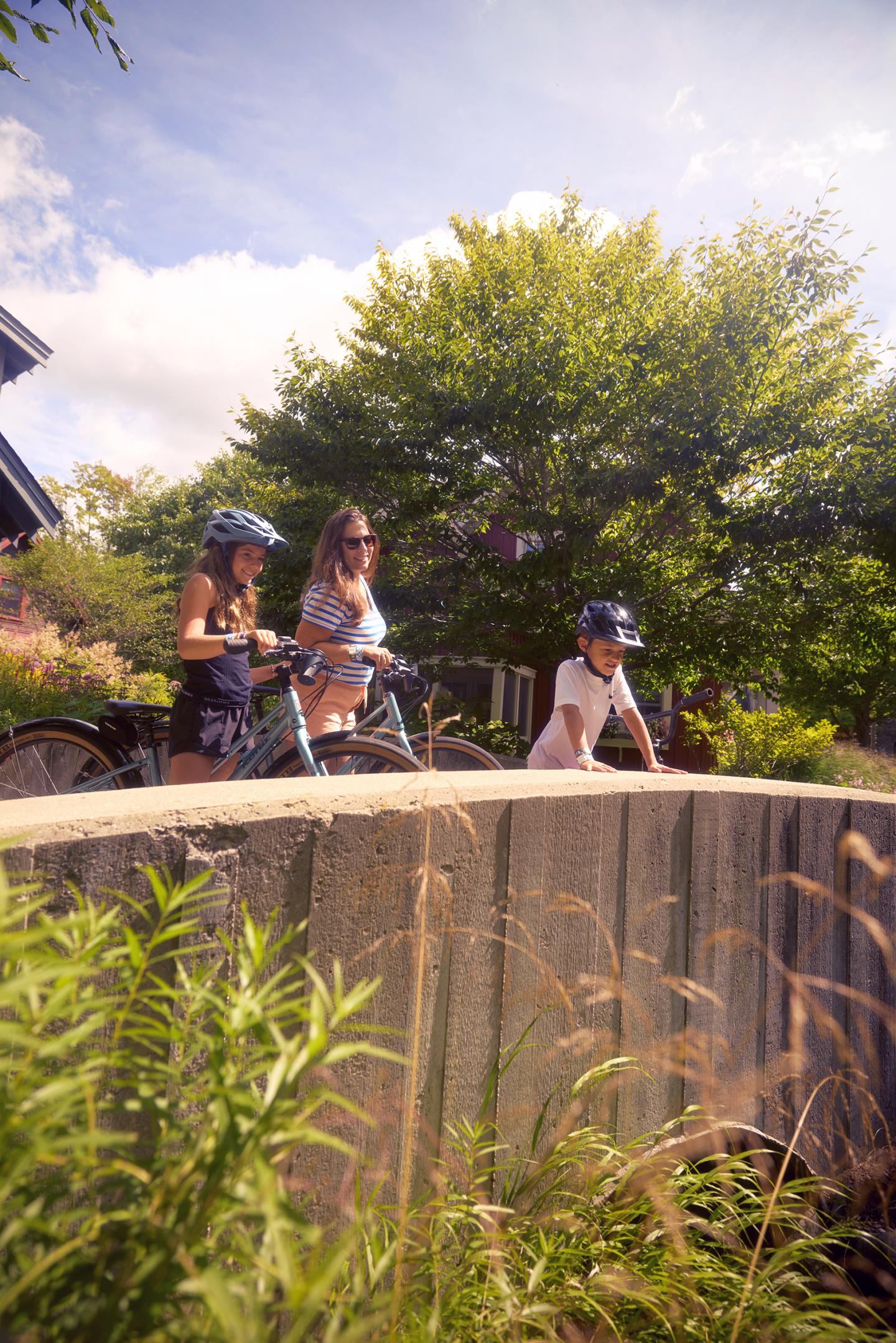 Three people with bikes and helmets near a concrete wall on a sunny day.