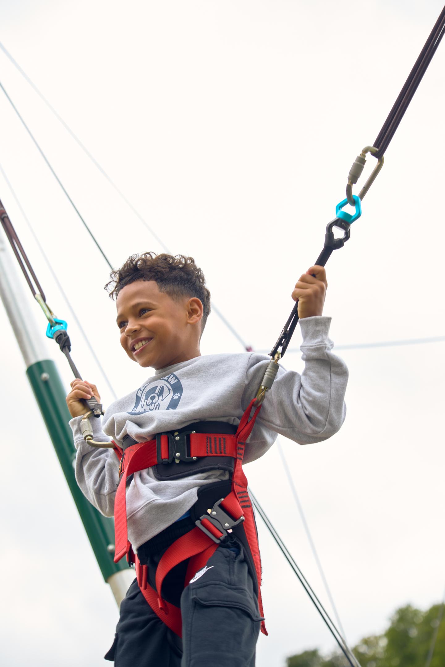 Child smiling while harnessed and holding bungee cords, sky in background.