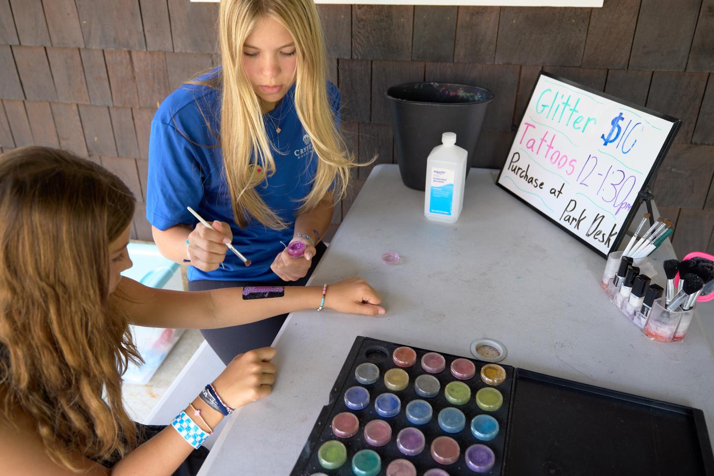 Two girls painting a temporary tattoo at an art booth with colorful paints.