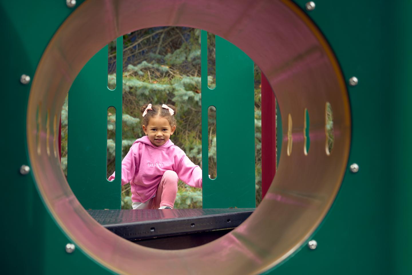 Toddler in pink jacket plays in a playground tunnel.