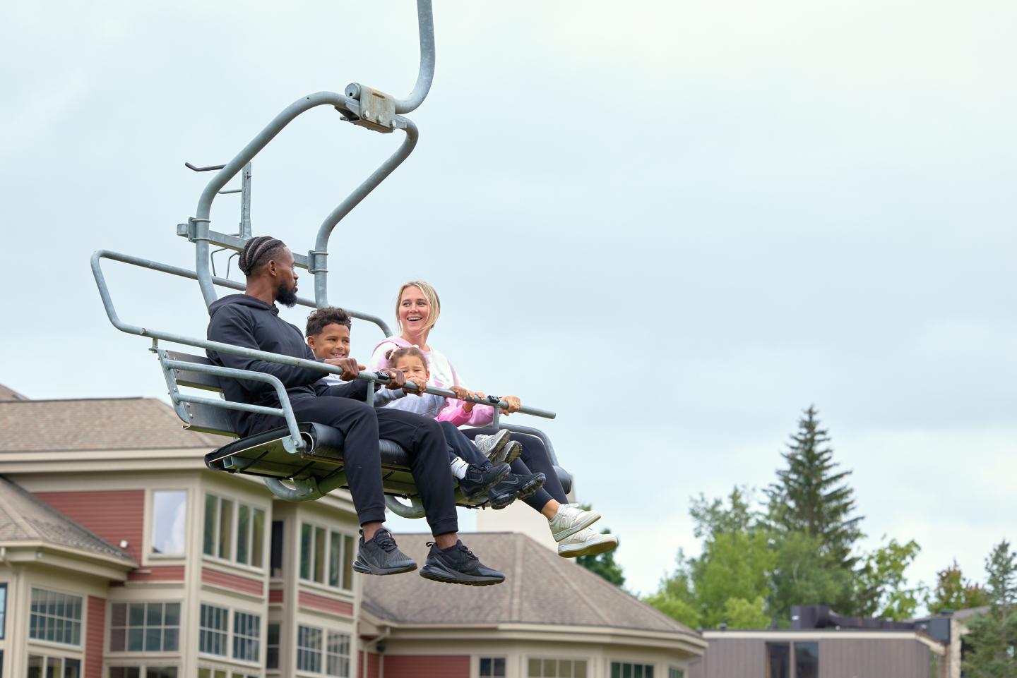 Four people smiling on a ski lift above buildings and trees.