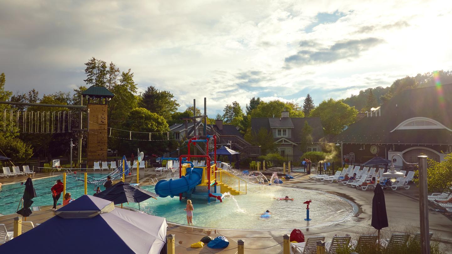 Outdoor pool with slides, surrounded by trees and lounge chairs under a cloudy sky.