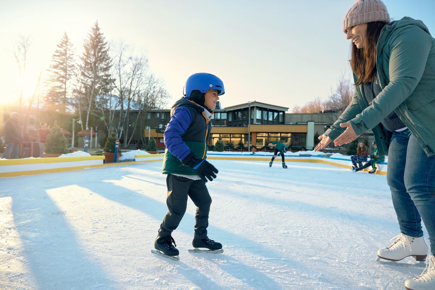 Child in helmet learning to ice skate, supported by an adult, on an outdoor rink at sunset.