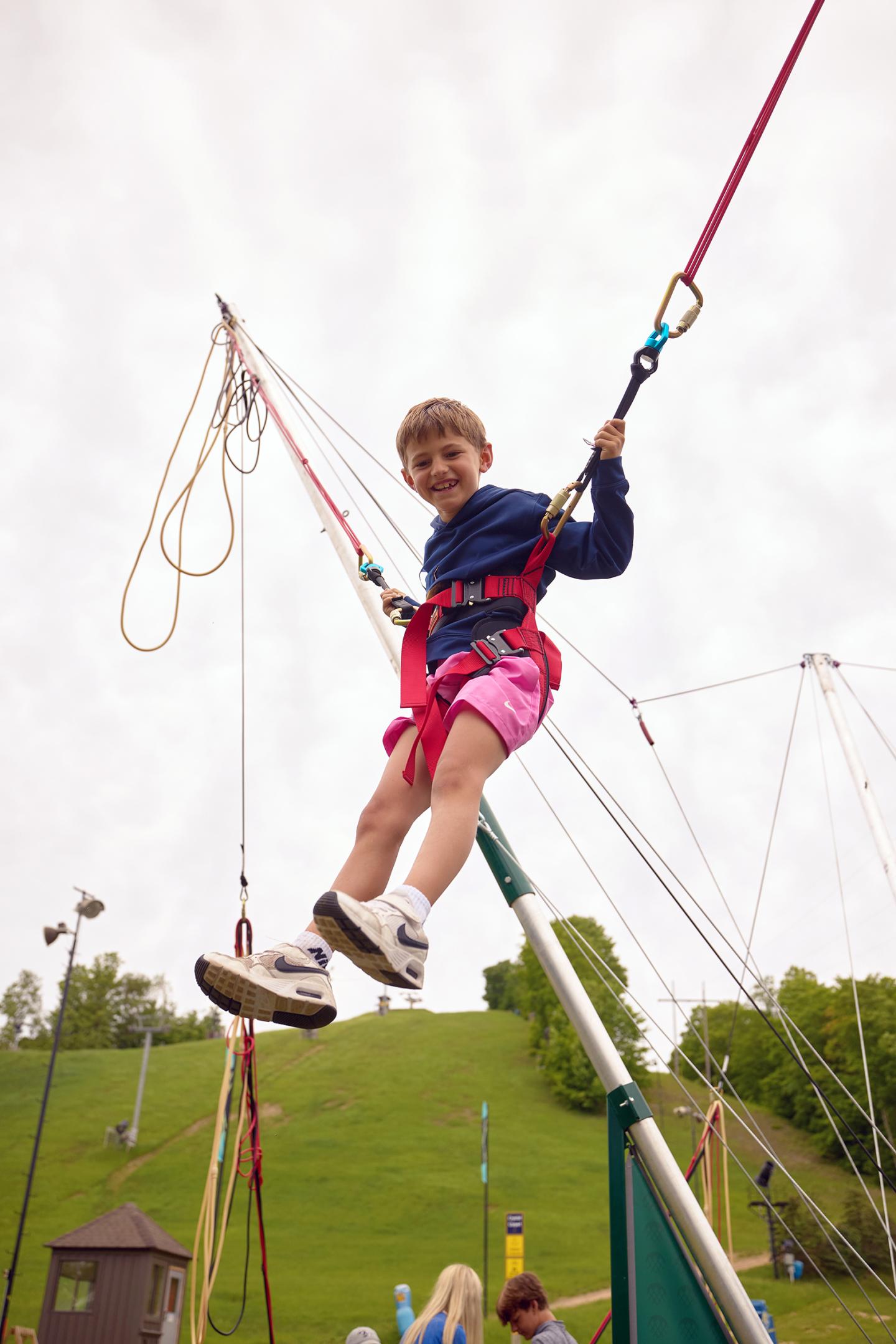 Boy smiling on a bungee trampoline, wearing pink shorts.