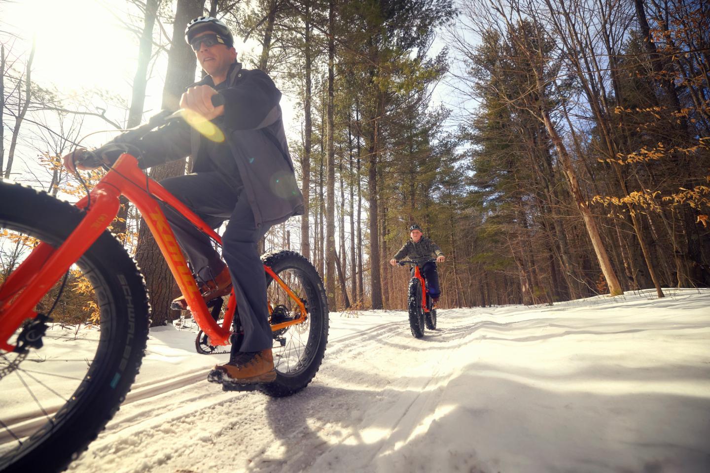Two people riding fat bikes on a snowy trail in a sunlit forest.