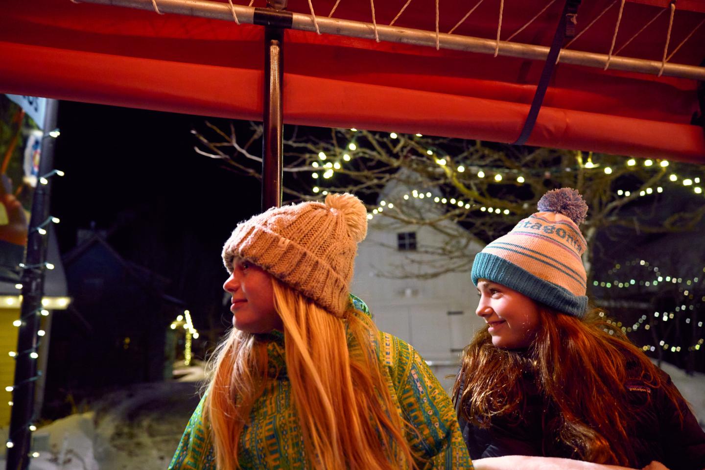 Two girls in winter hats, smiling under festive lights at night.