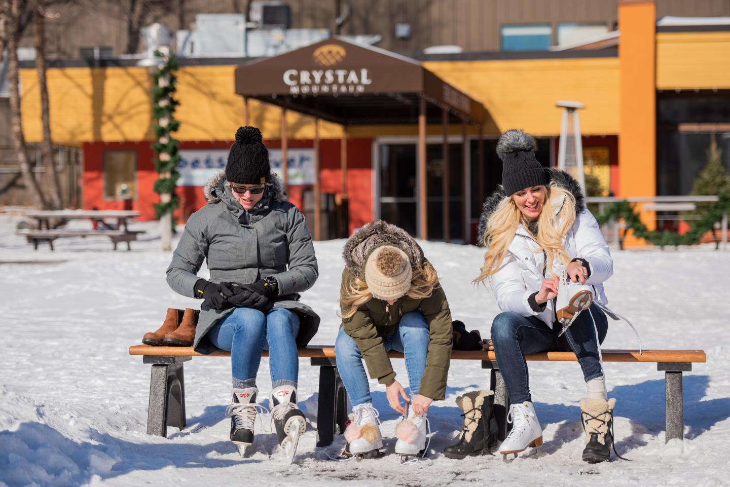 Three people sit on a bench in the snow, putting on ice skates.