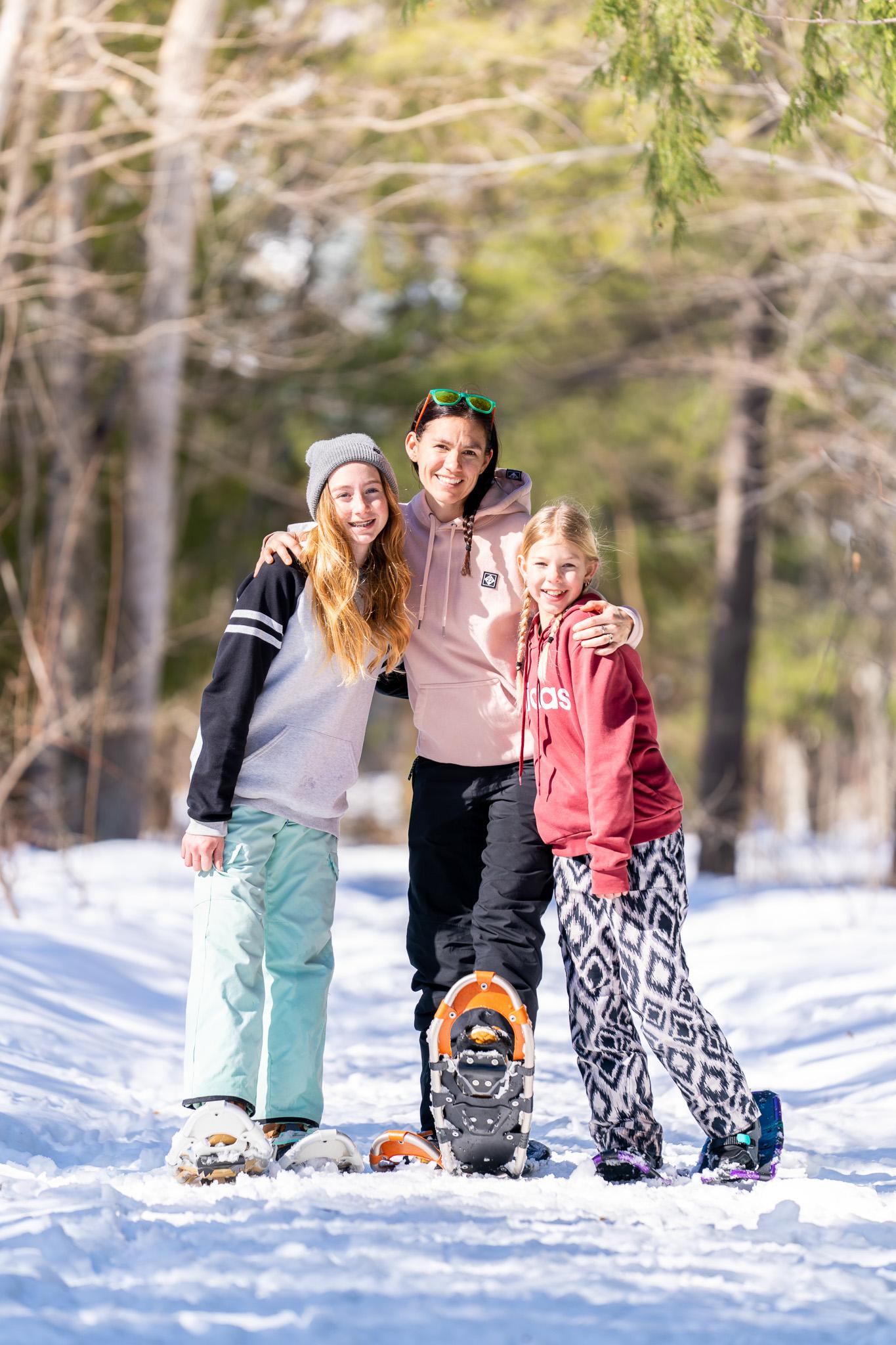 Three people smiling, standing on a snowy path, surrounded by trees.