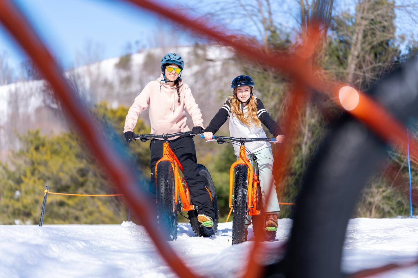 Two people biking on snow, framed by blurry bike wheels, wearing helmets.