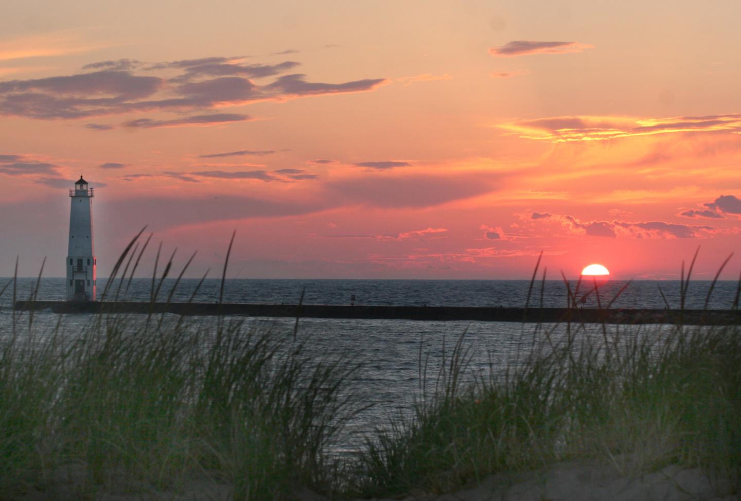 Sunset over the ocean with a distant lighthouse and grassy foreground.