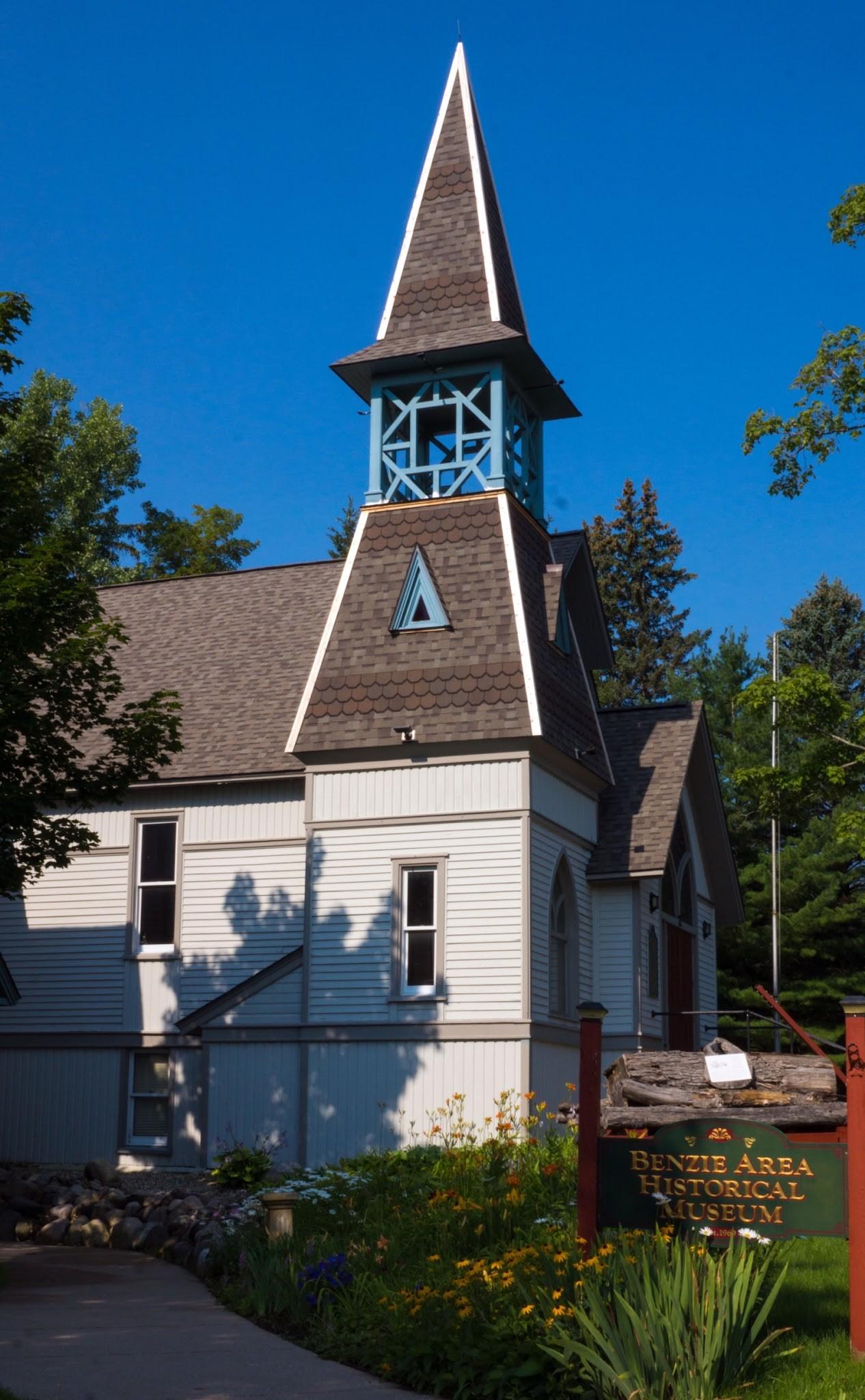 Church with a wooden steeple, surrounded by trees and flowers under a clear blue sky.