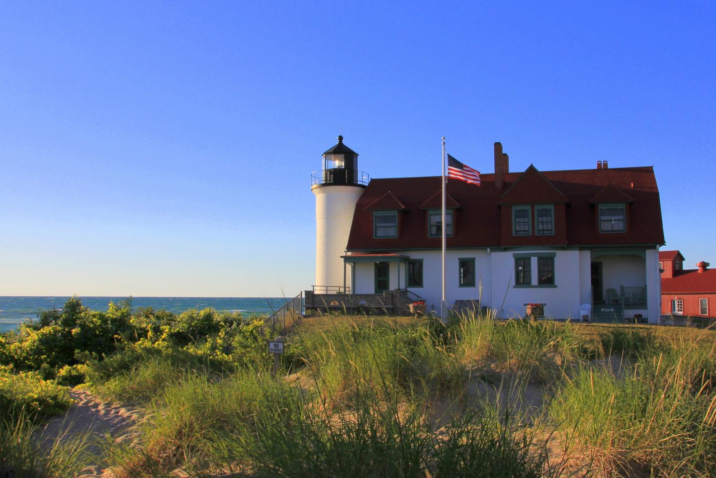 Coastal lighthouse at sunset, red-roofed building, grassy dunes in foreground.