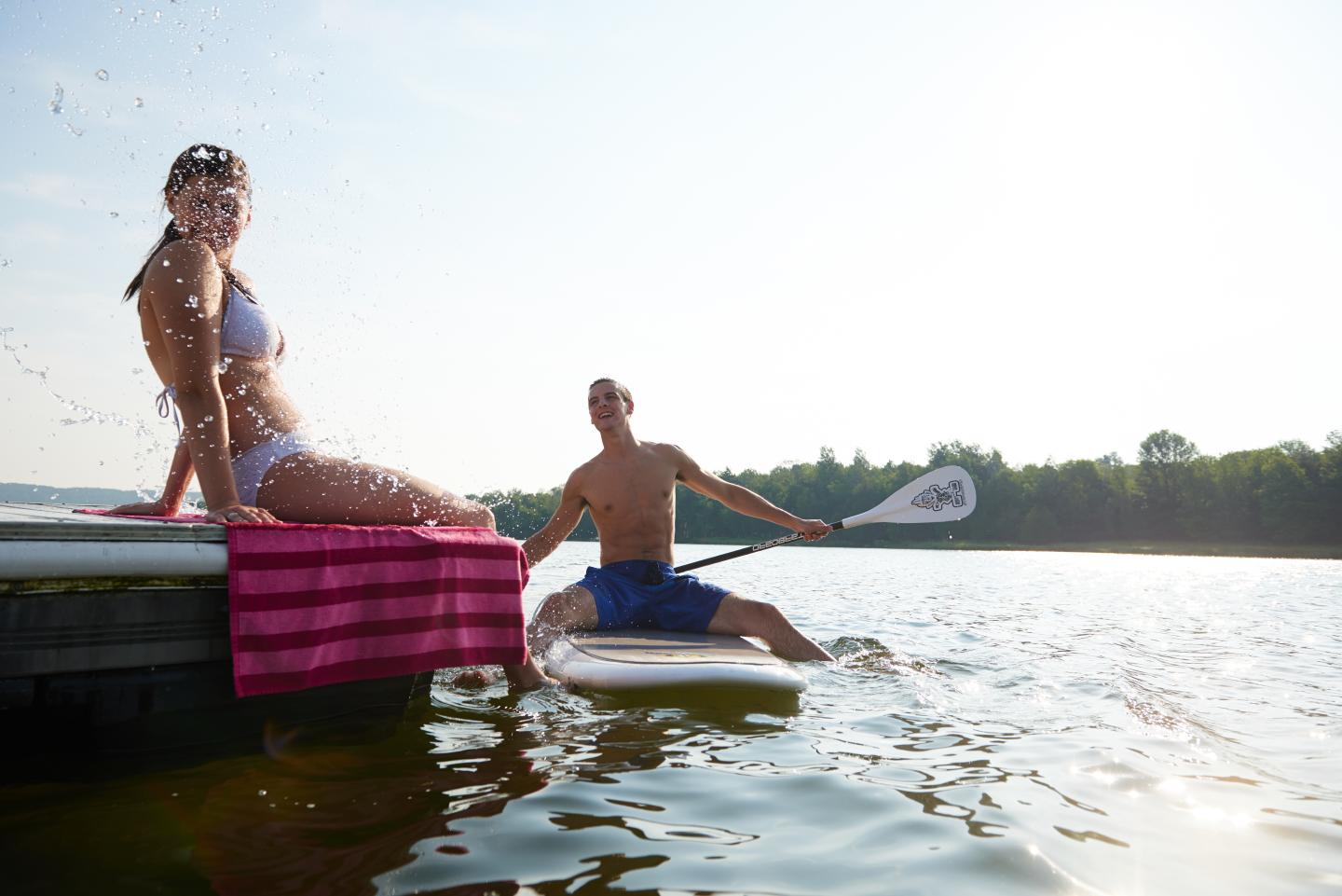 Man paddleboarding next to woman sitting on dock, both smiling under clear sky.
