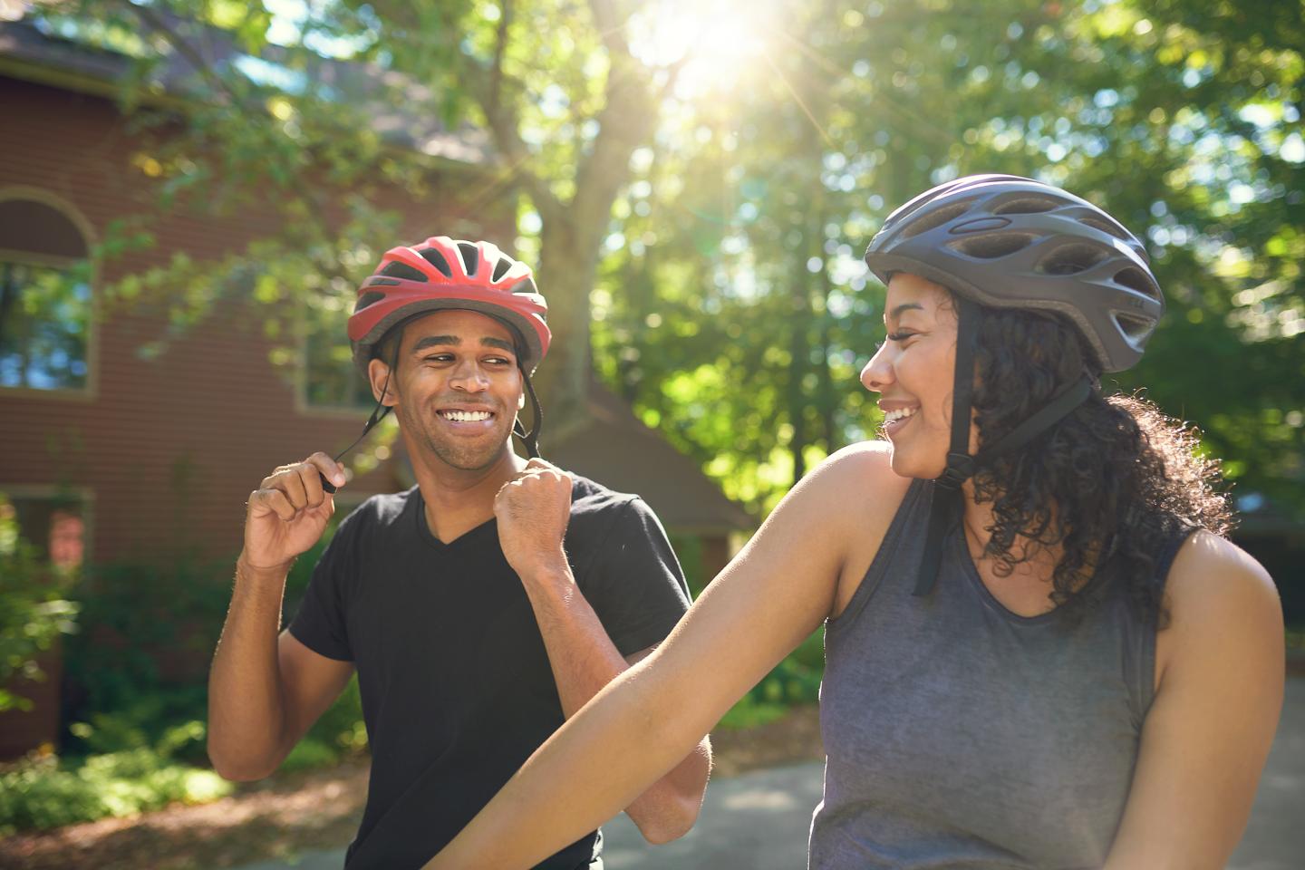 Two people wearing bike helmets, smiling under sunlight in a park setting.