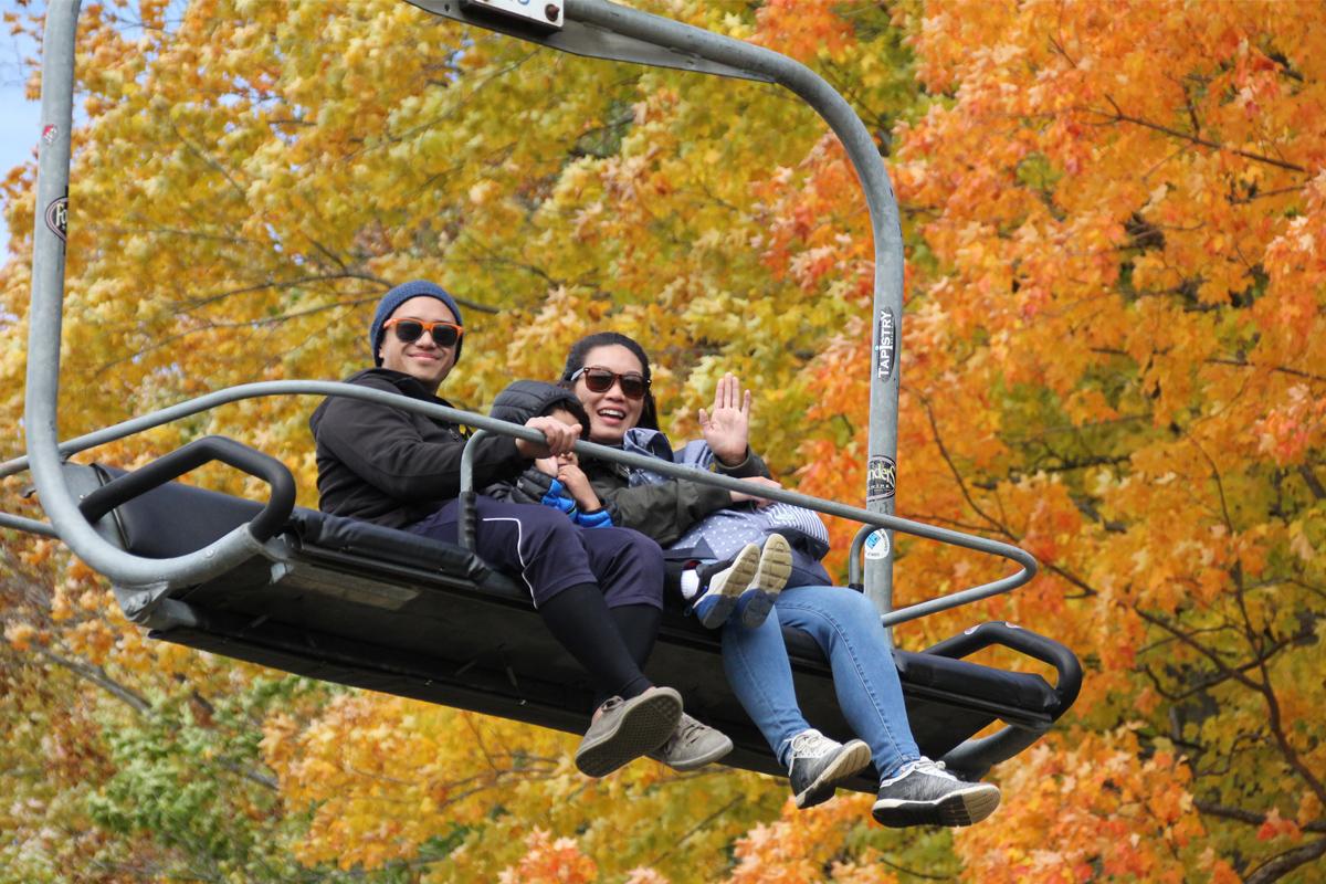 Three people on a ski lift, surrounded by autumn leaves, one waves.