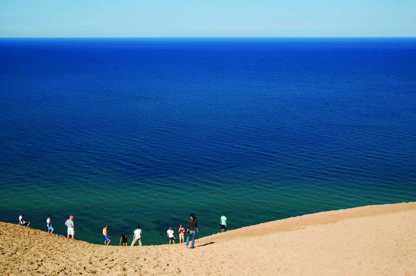 People walking down a sandy dune toward a vast blue lake.