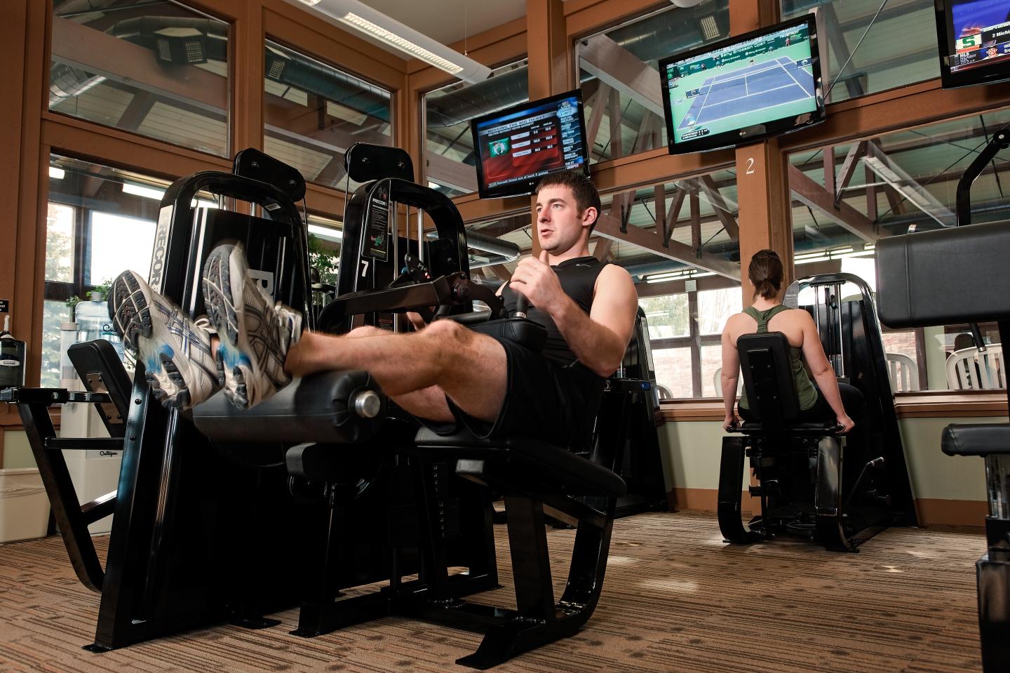 Man using leg press in a gym, with TV screens overhead.