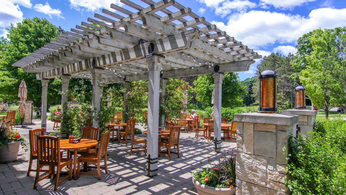 Outdoor patio with wooden furniture under a pergola, surrounded by lush greenery.