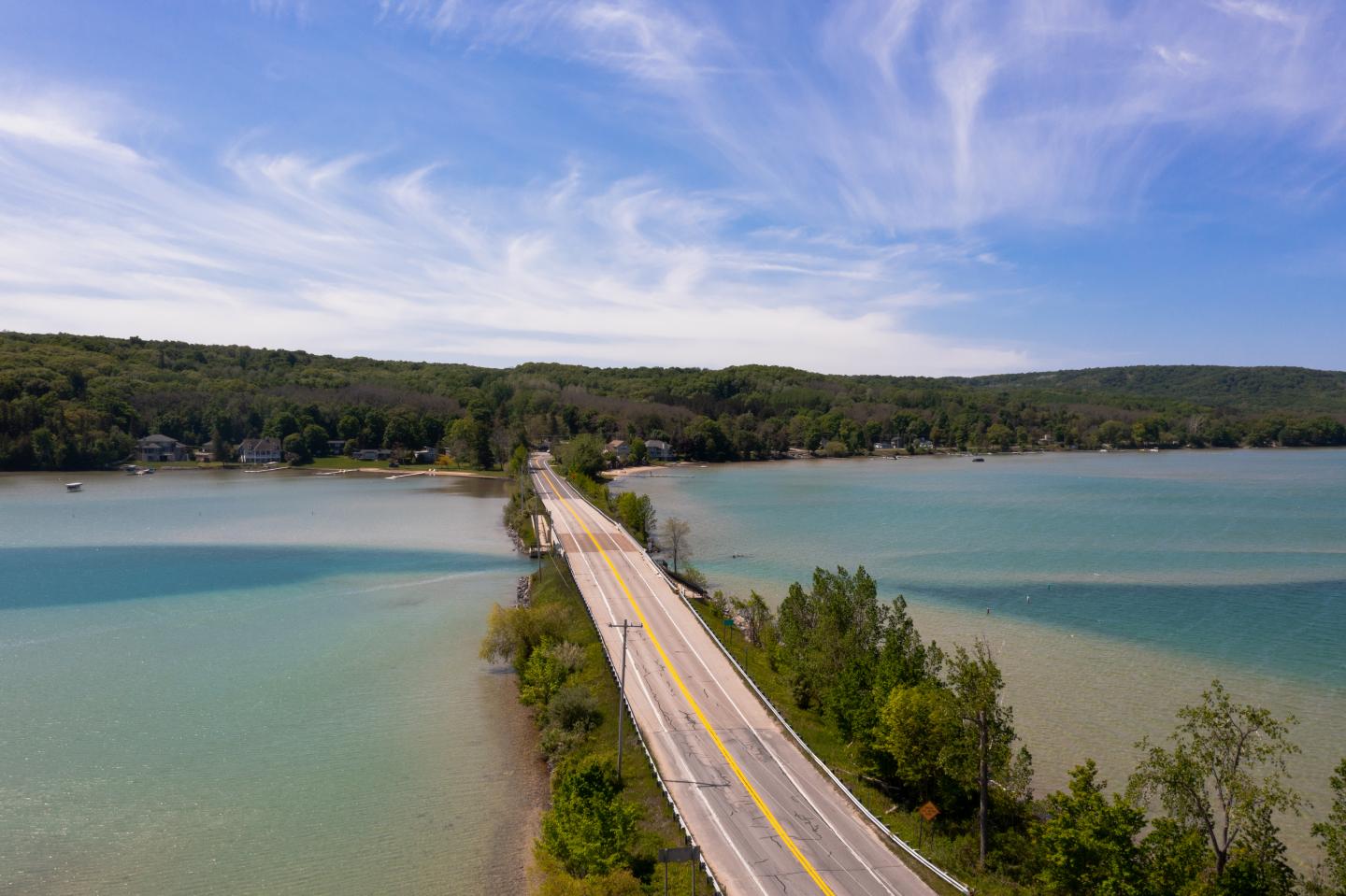 Aerial view of M-22 over a serene, blue-green lake with a forested shoreline.