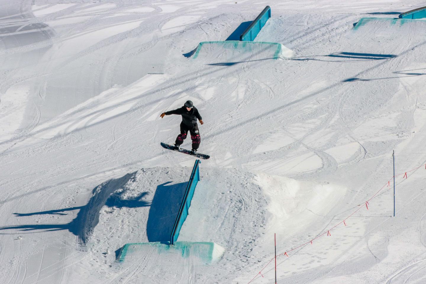 Snowboarder jumping in a snowy terrain park with ramps and rails.