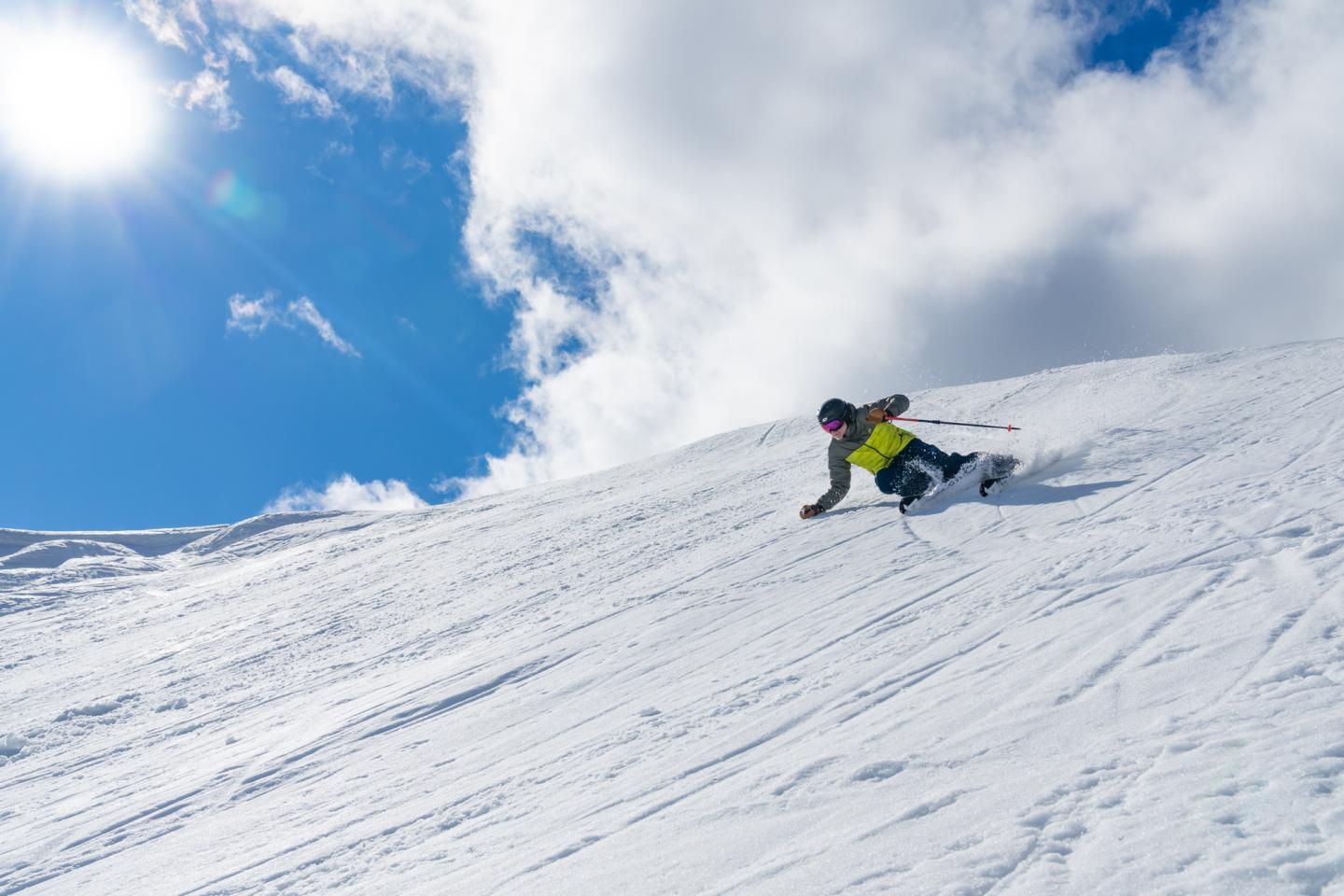 Skier in bright gear descends snowy slope under clear blue sky.