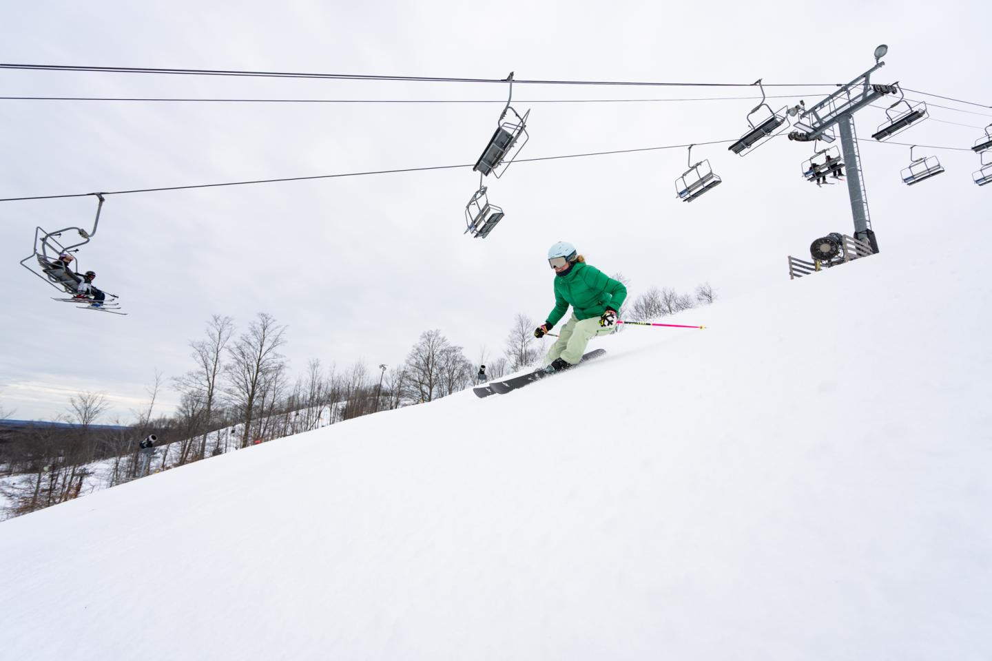 Skier in green jacket gliding down a snowy slope under ski lifts.