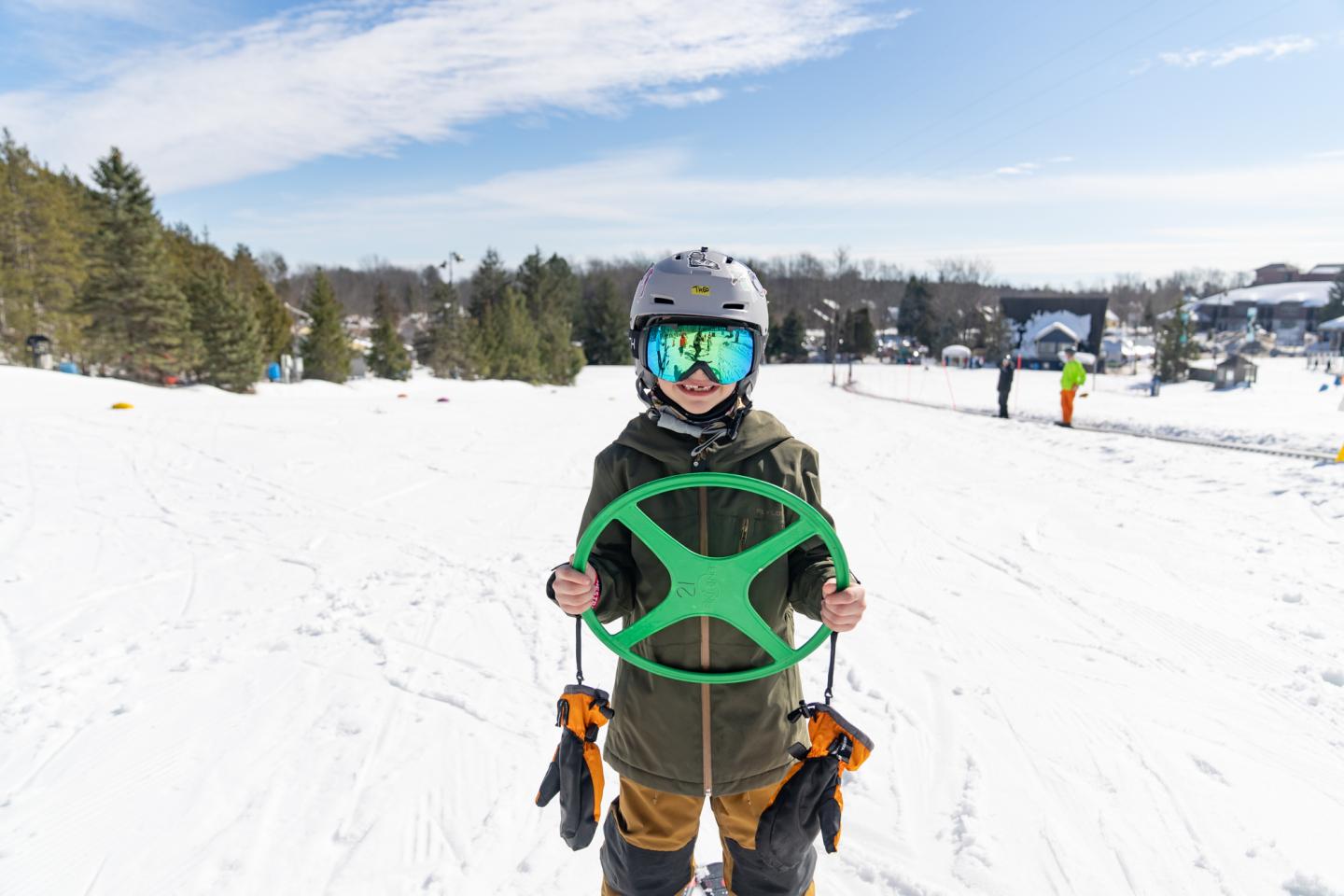 Child in snow gear holding green sled on a snowy hill.
