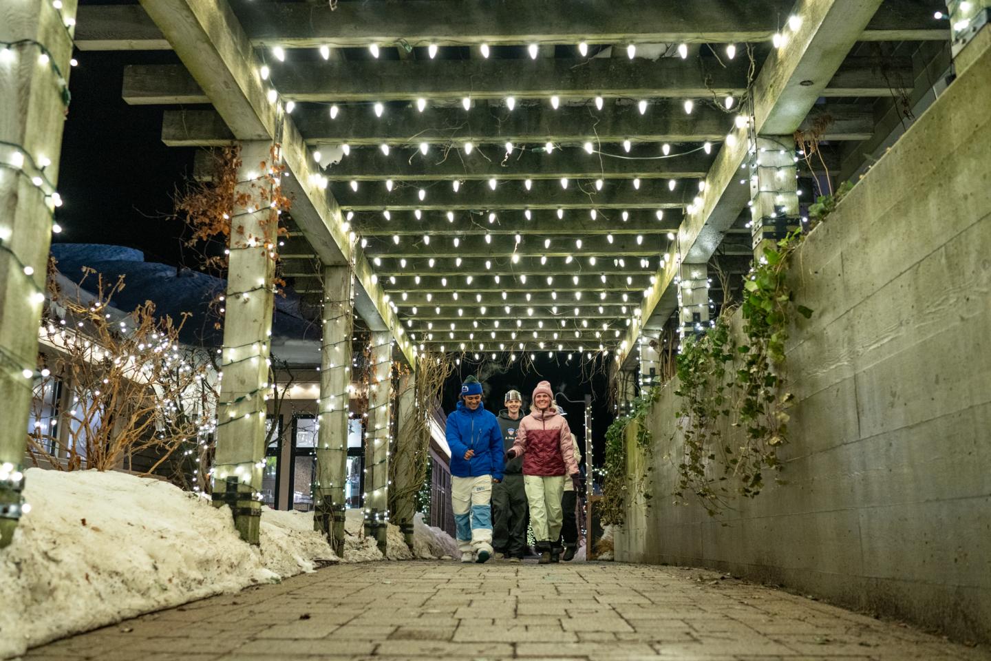 People walking under festive string lights on a snowy night.
