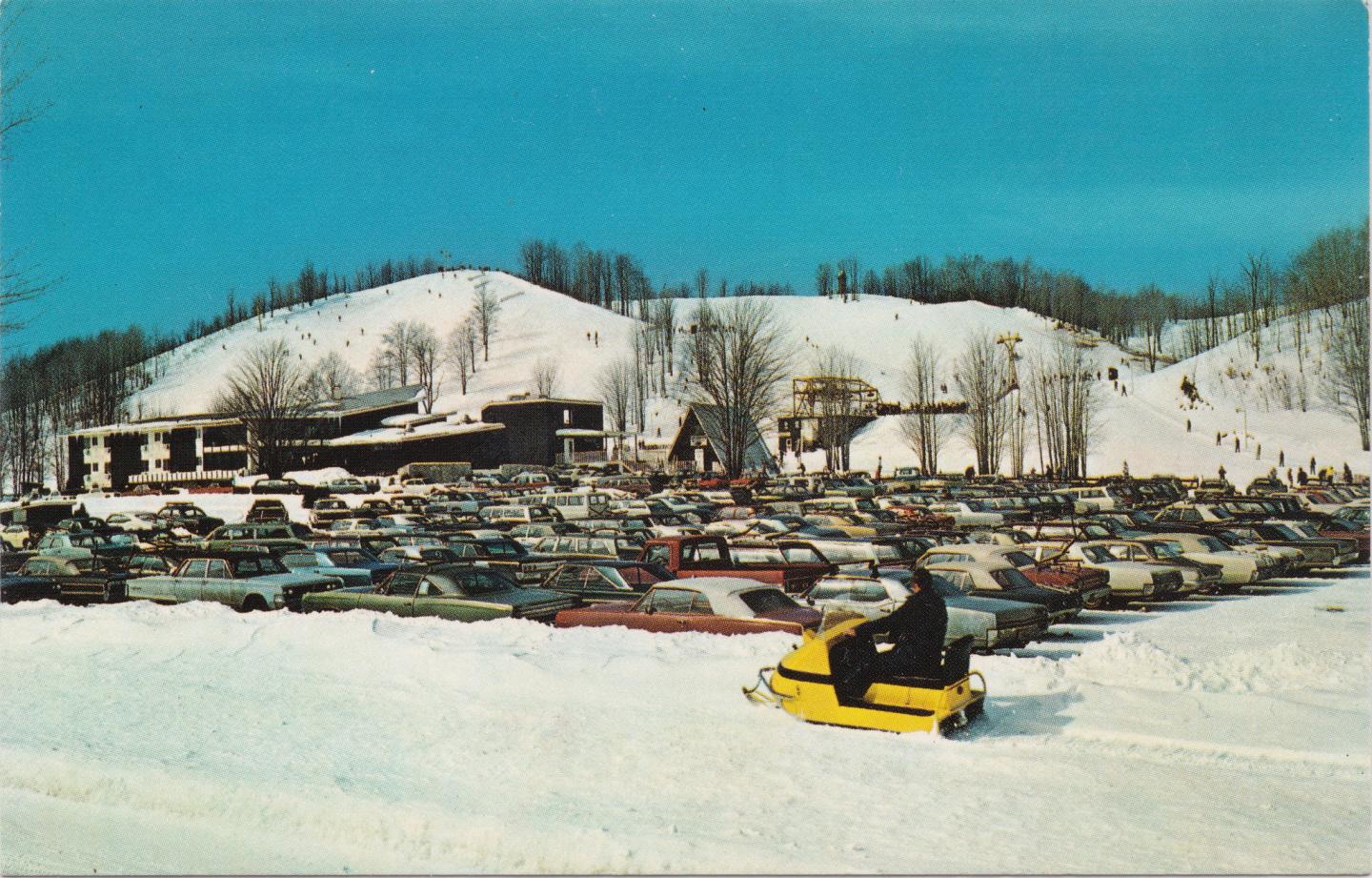 Snowy parking lot with many cars and a yellow snowmobile, trees and hills in the background.