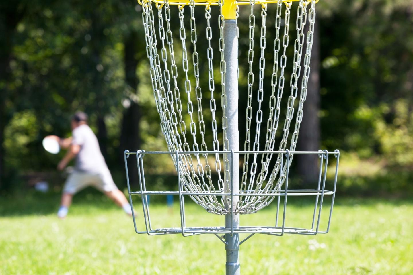 Disc golf basket with player in background, outdoors.