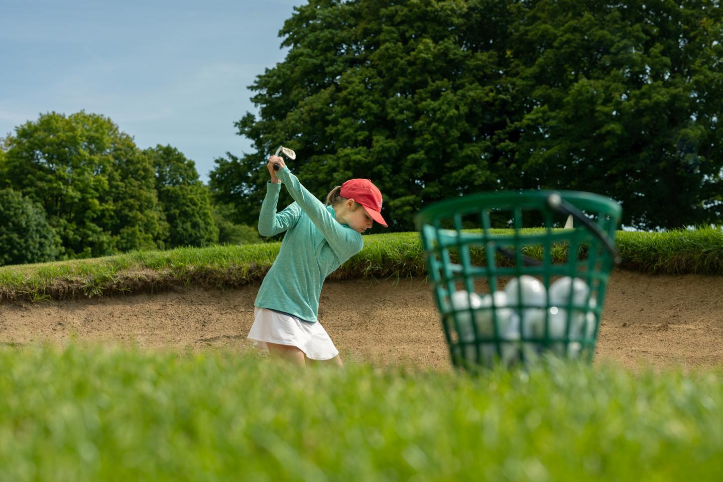 Child practicing golf swings near a basket of balls on a grassy course.