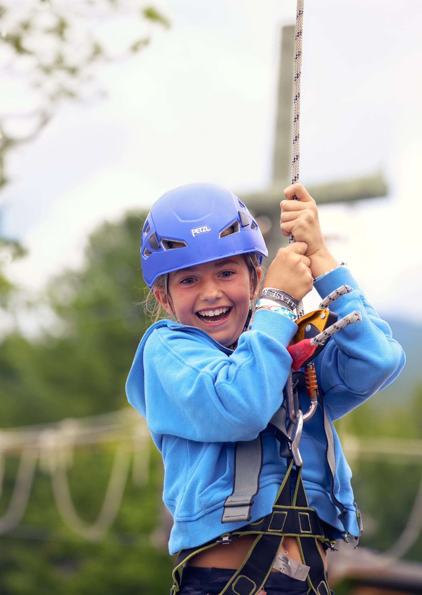 Young child zip-lining, wearing blue helmet and harness, smiling.