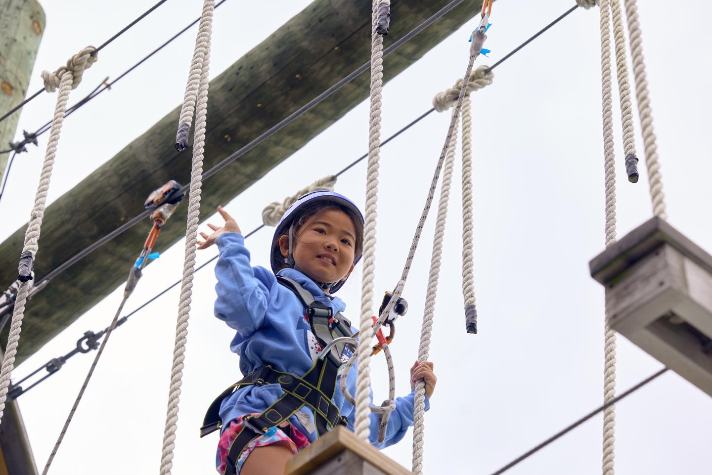 Child smiling on a ropes course, wearing a helmet and harness.