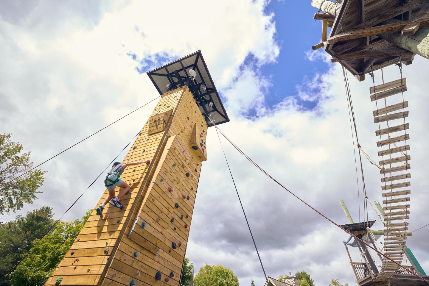 Climber scales a tall outdoor climbing wall under a partly cloudy sky.