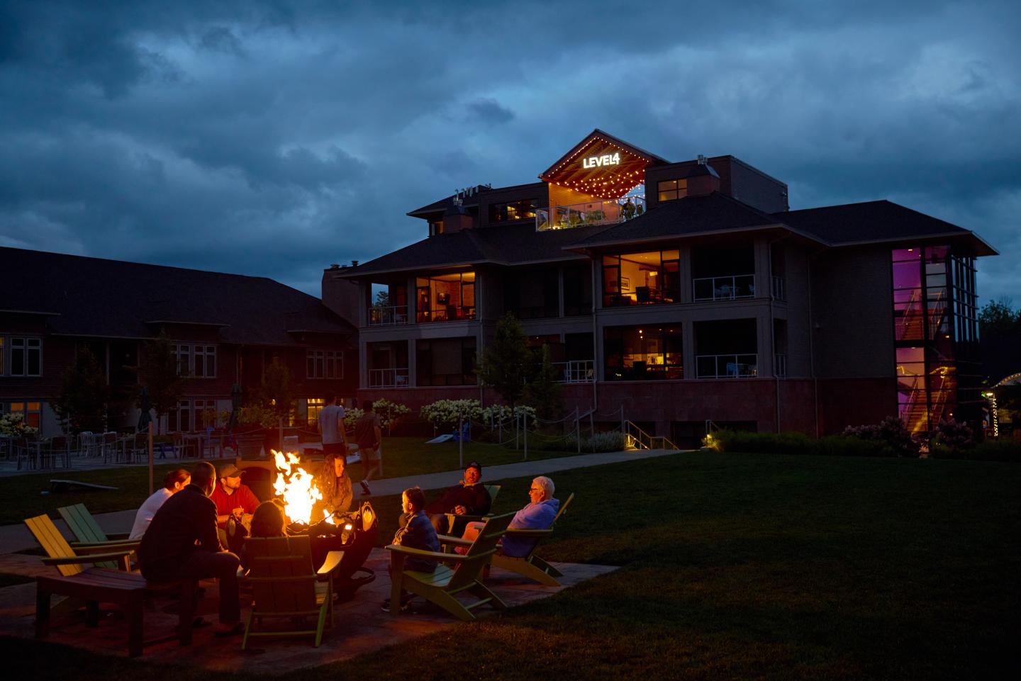 Group sitting around a campfire outside a large, illuminated building at dusk.