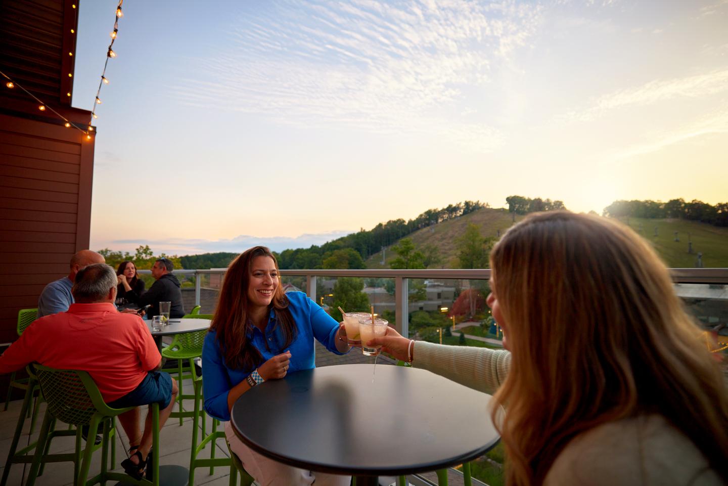Two people toasting at an outdoor table, sunset in the background.