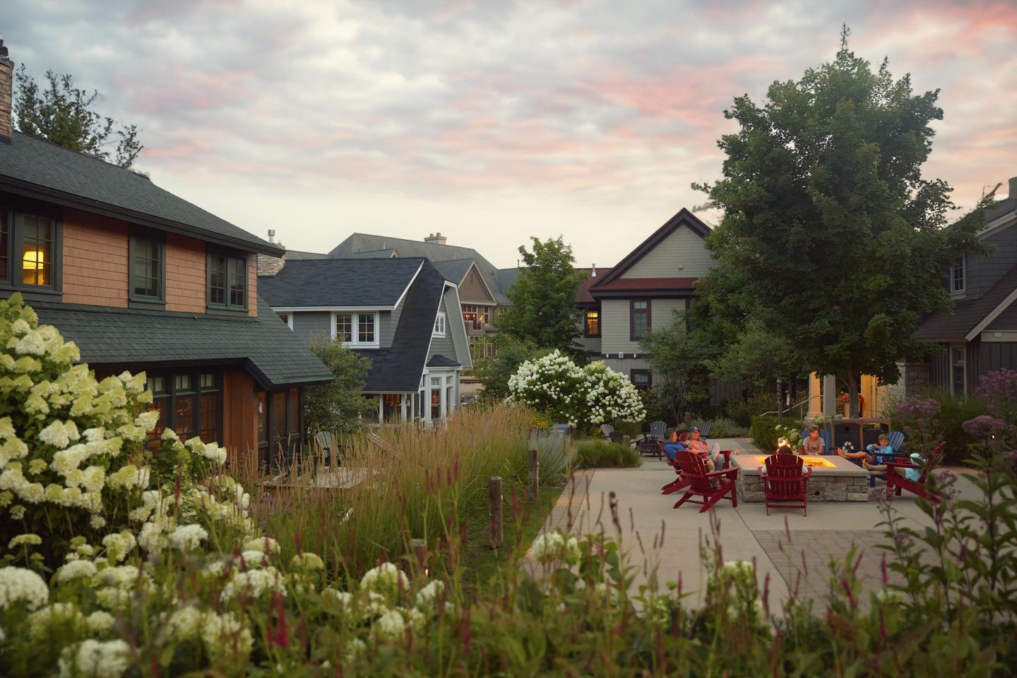 Courtyard with people sitting around a fire pit, surrounded by houses and flowers at sunset.