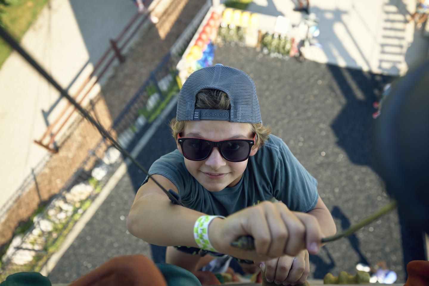 Boy climbing a wall, wearing sunglasses and a cap, in a sunny outdoor setting.