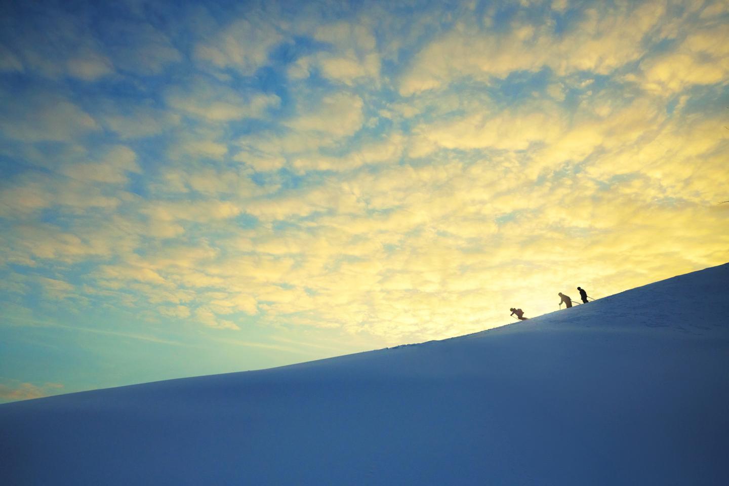 Silhouetted hikers ascend a snowy hill at sunrise under a colorful sky.