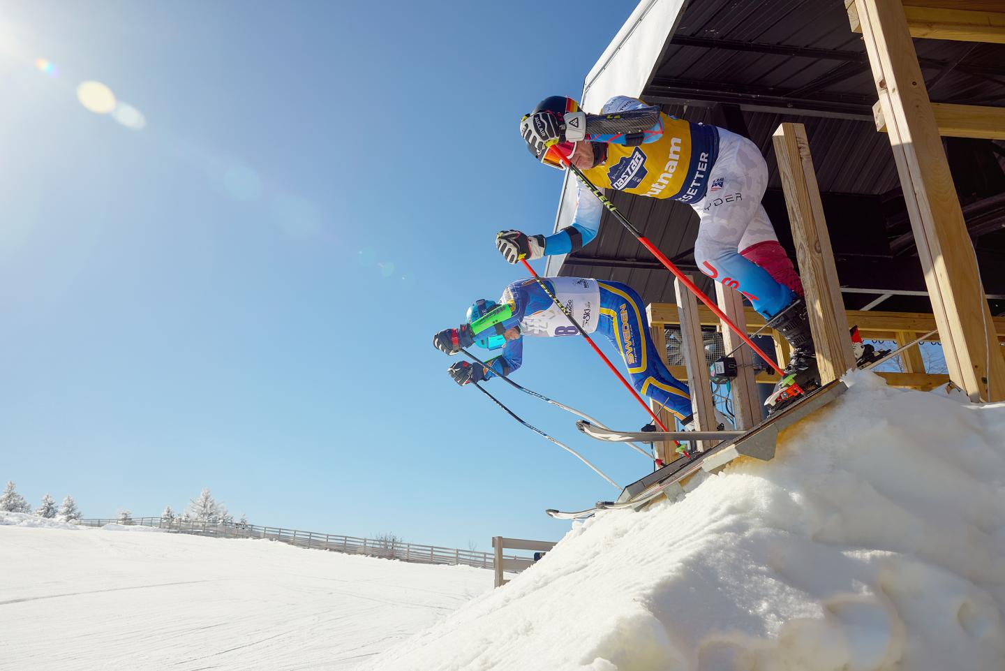 Skiers race downhill from a snowy start gate under a clear blue sky.
