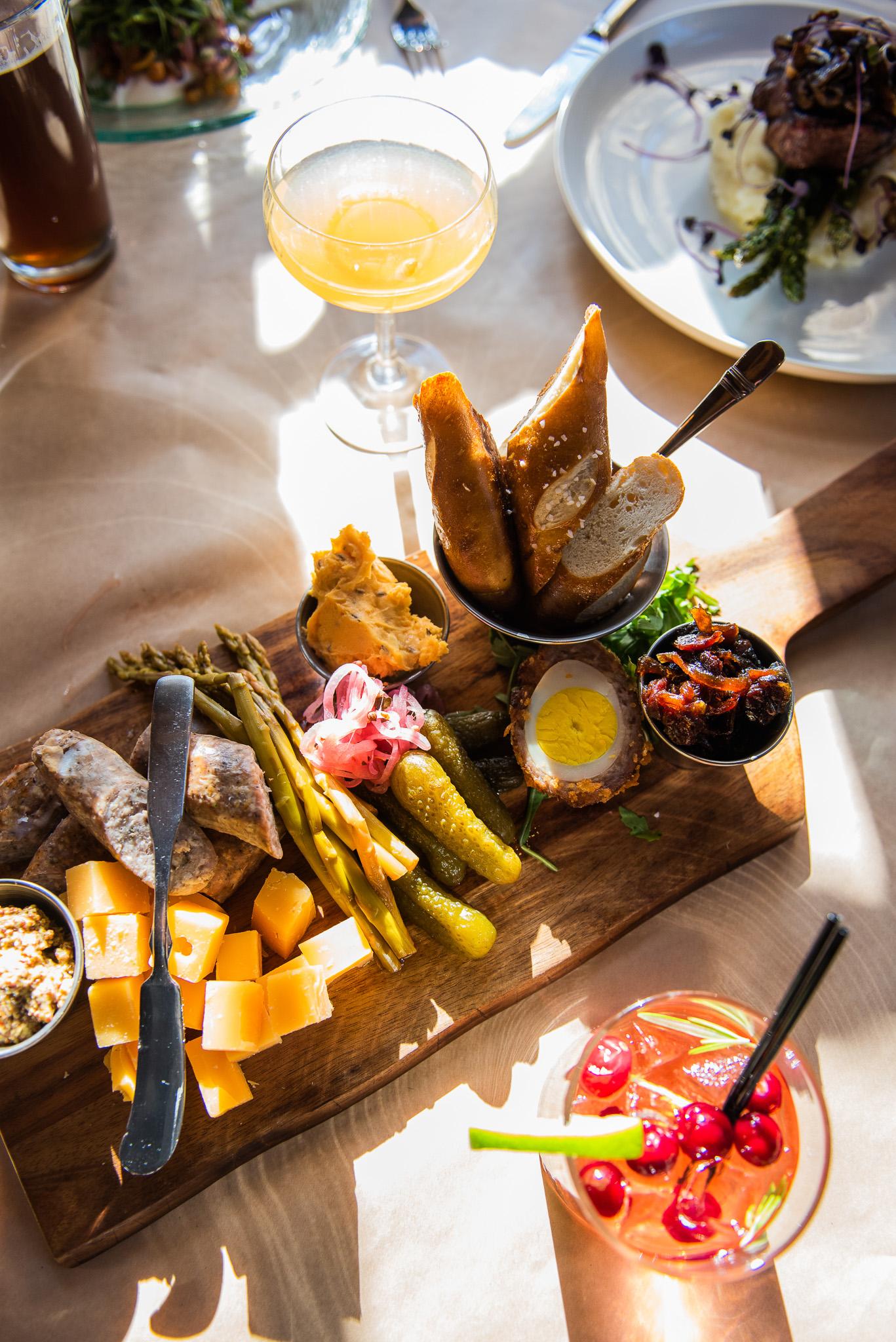 Charcuterie board with cheeses, meats, pickles, and a cocktail, in a sunlit setting.